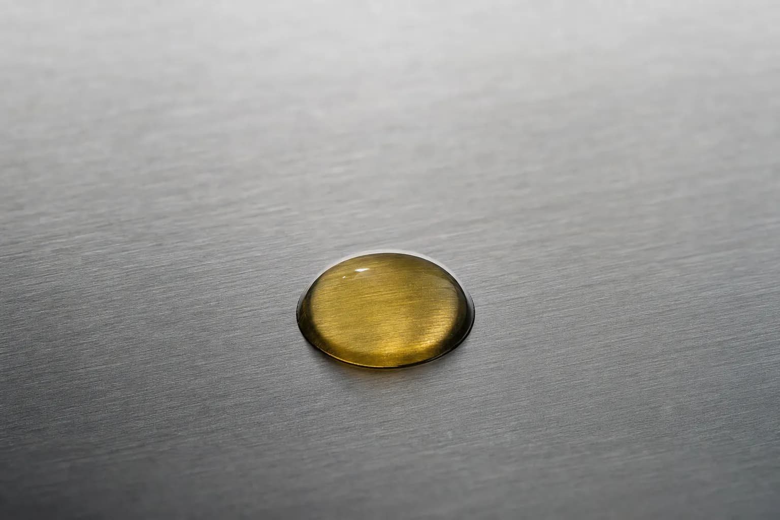 Macro photograph of a single translucent amber droplet of marine fish oil resting on a brushed-stainless-steel laboratory bench surface under cold Nordic daylight